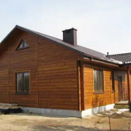 Gotowy dom z drewna o powierzchni 96 m² z brązowym dachem i kominem, widok z boku na tle zimowego krajobrazu z resztkami śniegu.